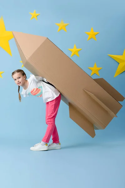 Cute kid in pink pants holding big cardboard rocket on blue starry background — Stock Photo