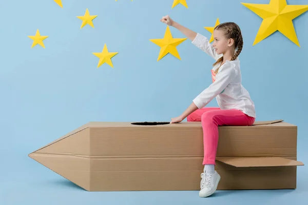 Preteen enfant assis sur une fusée en carton avec poing vers le haut sur fond bleu avec des étoiles — Photo de stock