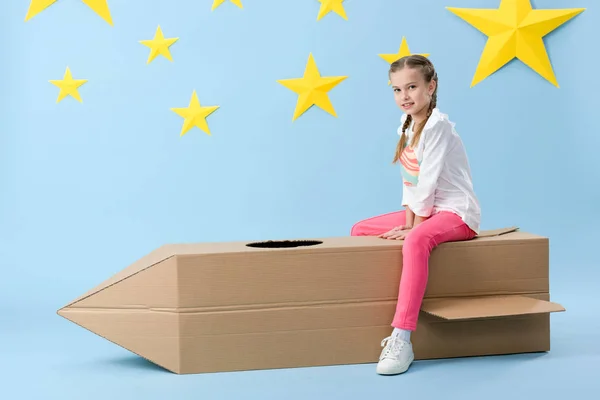 Улыбающийся ребенок с косичками сидит на картонной ракете на синем звездном фоне — стоковое фото