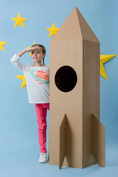 Веселый ребенок стоит рядом с картонной ракетой и смотрит в сторону на голубом звездном фоне — стоковое фото