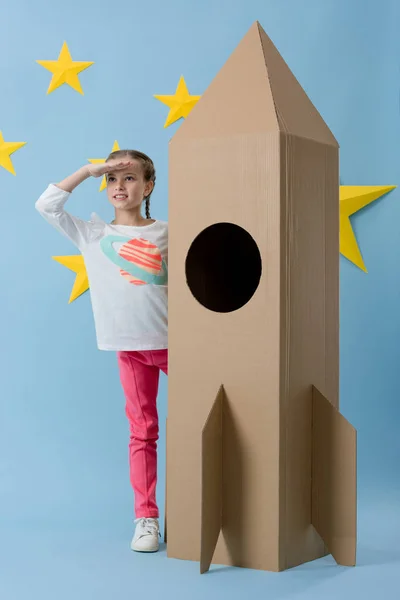 Alegre niño de pie cerca de cohete de cartón y mirando hacia otro lado sobre fondo azul estrellado - foto de stock