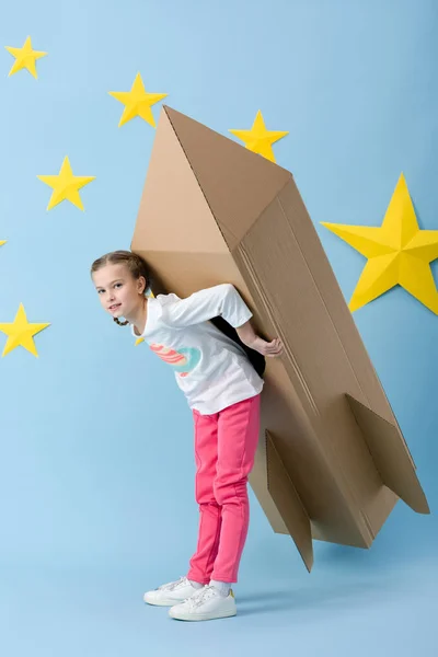 Enfant insouciant en pantalon rose tenant une grosse fusée en carton sur fond bleu avec des étoiles — Photo de stock