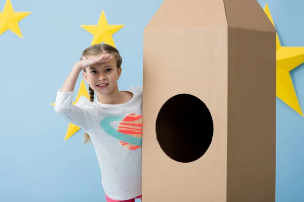 Sonriente niño de pie cerca de cohete de cartón y mirando hacia otro lado sobre fondo azul estrellado - foto de stock