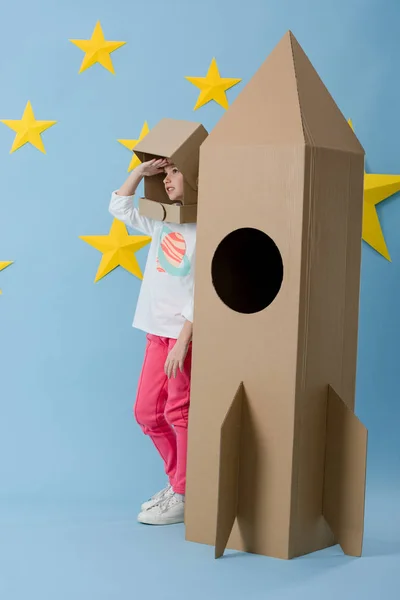 Kid in cardboard helmet standing near rocket and looking away on blue starry background — Stock Photo