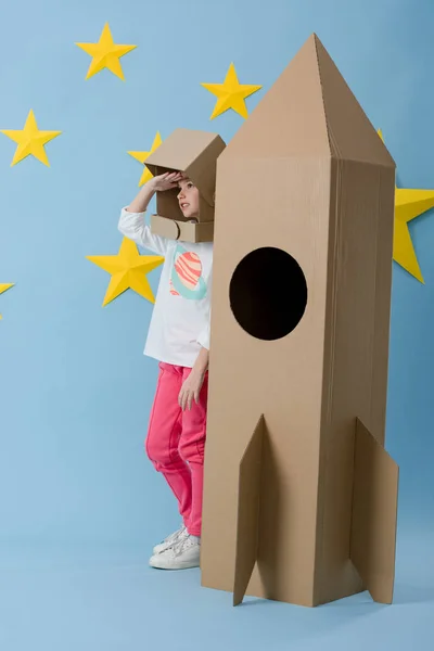 Enfant en carton casque debout près de la fusée et regardant loin sur fond bleu étoilé — Photo de stock