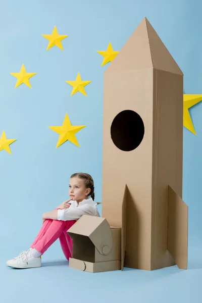 Dreamy kid with helmet sitting near cardboard rocket on blue starry background — Stock Photo