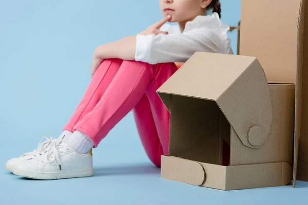 Cropped view of pensive child sitting with cardboard helmet on blue background — Stock Photo