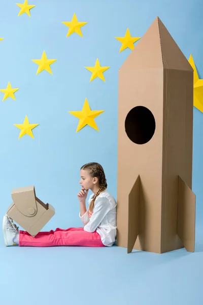 Side view of pensive kid sitting near cardboard rocket and looking at helmet on blue starry background — Stock Photo