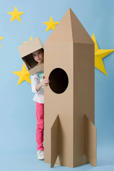 Full length view of kid in helmet standing near cardboard rocket on blue starry background — Stock Photo