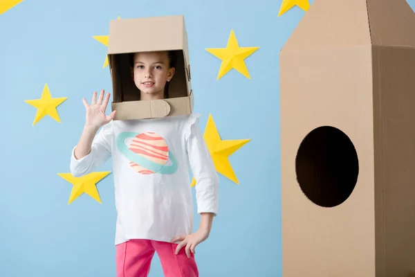 Enfant mignon dans un casque debout près de fusée en carton et agitant la main sur fond bleu étoilé — Photo de stock