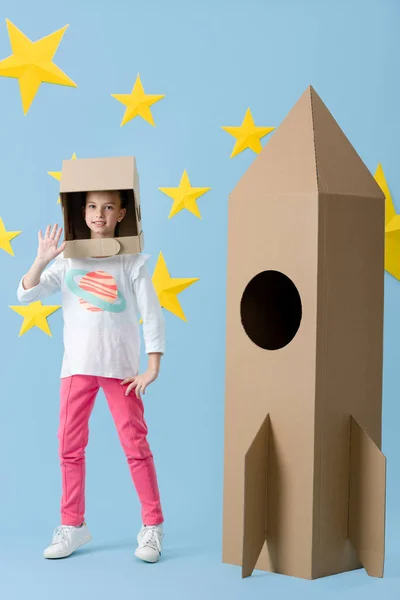 Щаслива дитина в шоломі стоїть біля картонної ракети і махає рукою на синьому фоні з зірками — стокове фото