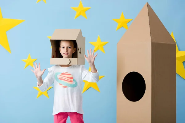 Interested kid in helmet waving hands near cardboard rocket on blue starry background — Stock Photo