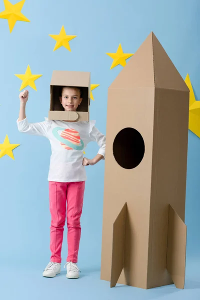 Criança inspirada no capacete gesticulando perto de foguete de papelão no fundo estrelado azul — Fotografia de Stock