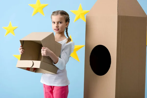 Kid with braids standing near cardboard rocket and holding helmet on blue starry background — Stock Photo