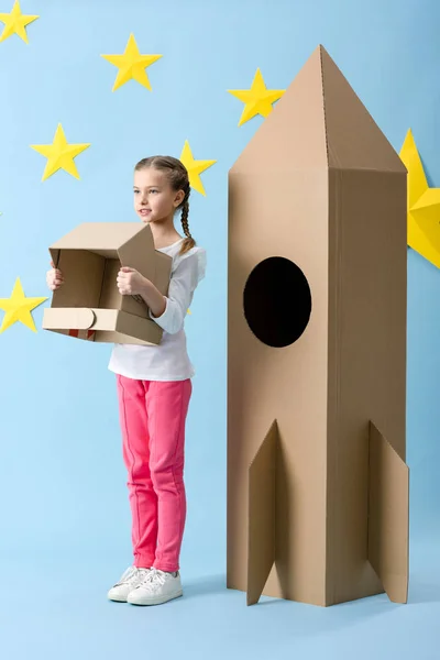 Inspired kid standing near big cardboard rocket and holding helmet on blue starry background — Stock Photo