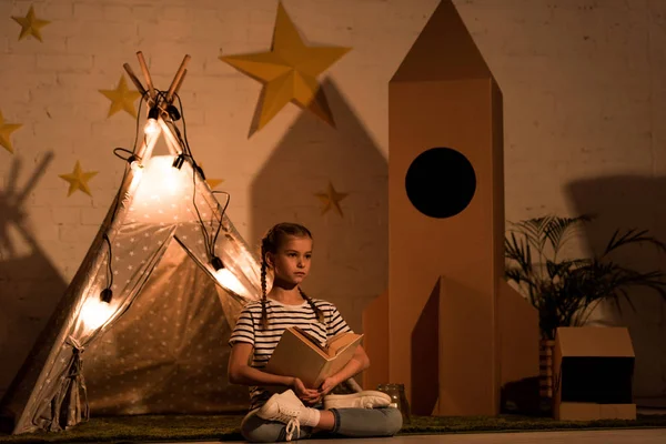 Relajado niño sentado en pose de loto y libro de lectura en habitación oscura - foto de stock