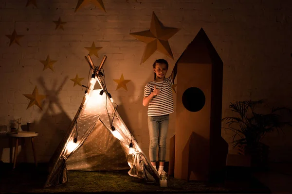 Criança de pé entre wigwam e foguete de papelão no quarto escuro — Fotografia de Stock