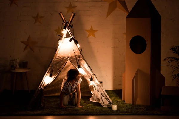 Kid standing on all fours in wigwam and looking at cardboard rocket in dark room — Stock Photo