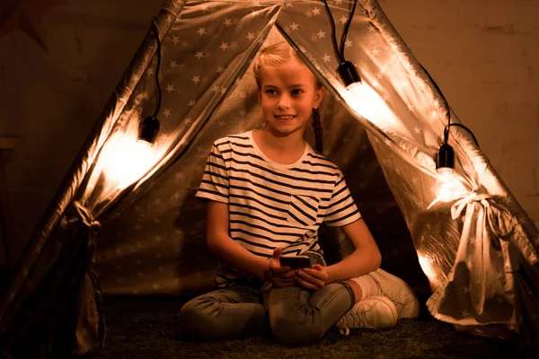 Счастливый ребенок со смартфоном сидит в уютном вигваме дома — стоковое фото