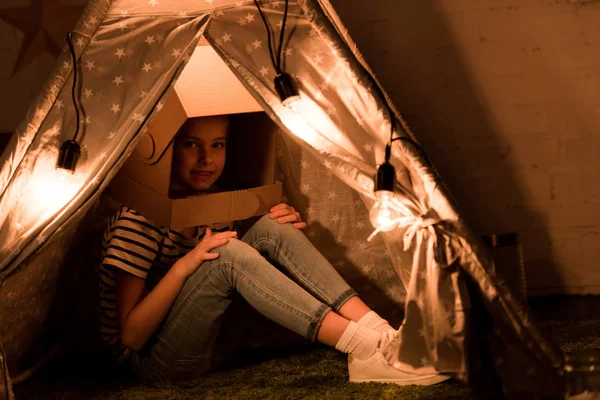 Bambino carino in casco di cartone seduto in wigwam con lampadine in camera oscura — Foto stock