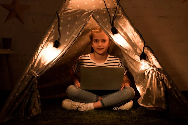 Happy kid using laptop while sitting in wigwam in dark room — Stock Photo