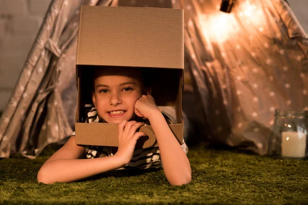 Carefree kid in cardboard helmet lying on carpet and looking at camera — Stock Photo