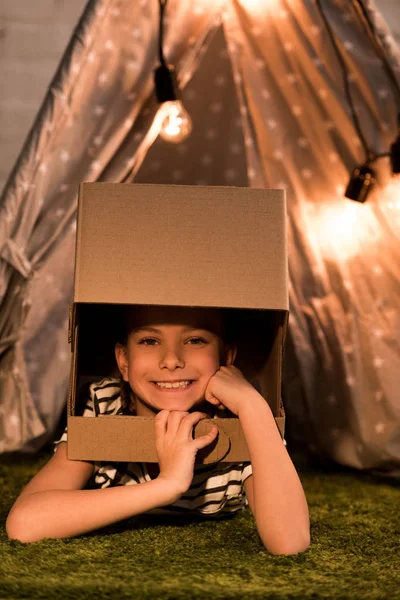 Laughing kid in cardboard helmet lying on green carpet — Stock Photo