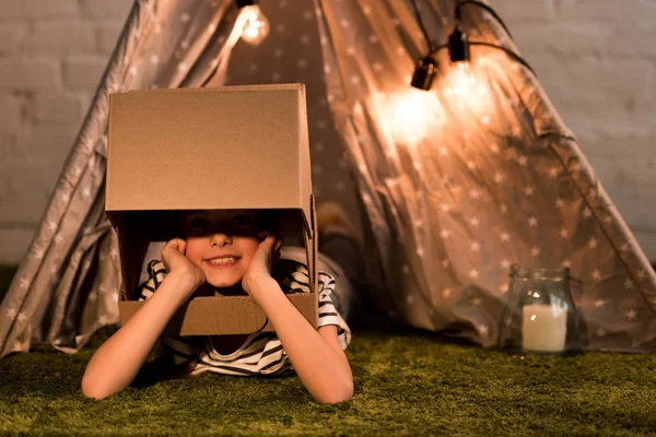Enfant drôle dans un casque en carton couché sur un tapis vert — Photo de stock