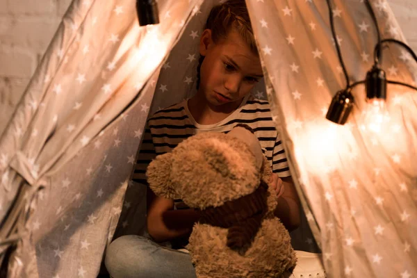 Disappointed kid looking at teddy bear while sitting in wigwam at home — Stock Photo