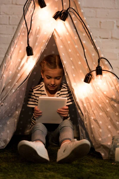 Улыбающийся ребенок сидит в уютном вигваме и держит цифровой планшет — стоковое фото