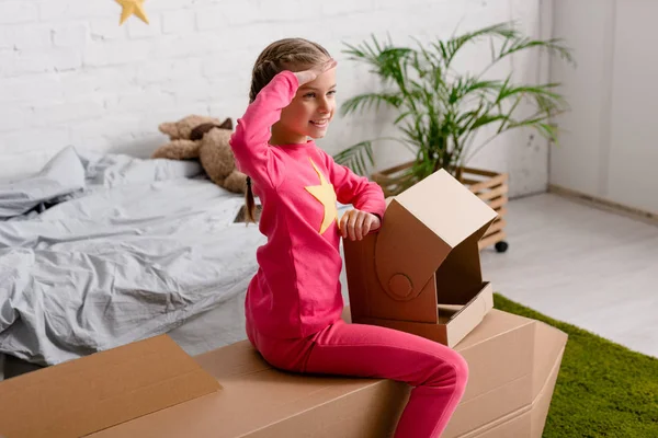 Lächelndes Kind mit Helm grüßt, während es im Schlafzimmer auf einer Papprakete sitzt — Stockfoto