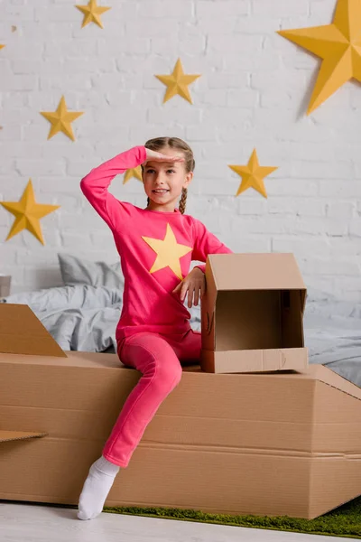 Excited kid sitting on cardboard rocket and looking away with smile — Stock Photo