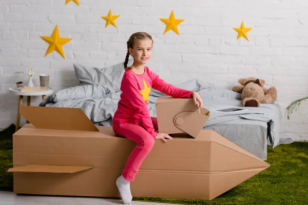 Enfant joyeux avec casque assis sur une fusée en carton dans la chambre — Photo de stock