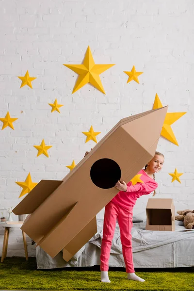 Visão de comprimento total da criança animada segurando grande foguete de papelão no quarto — Fotografia de Stock