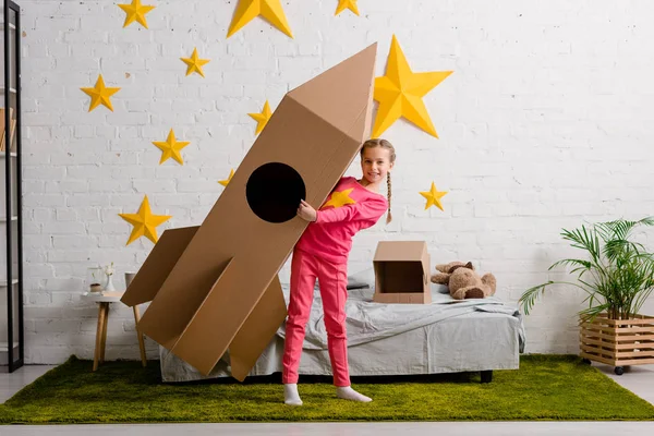 Niño dichoso en ropa rosa sosteniendo cohete de cartón grande en el dormitorio - foto de stock