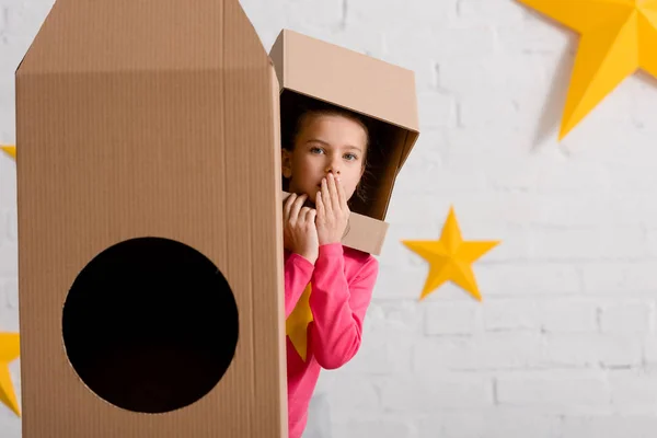 Surprised kid in cardboard helmet standing near rocket and covering mouth with hand — Stock Photo