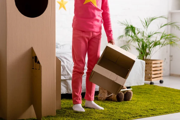 Cropped view of kid holding cardboard helmet and standing near rocket in bedroom — Stock Photo