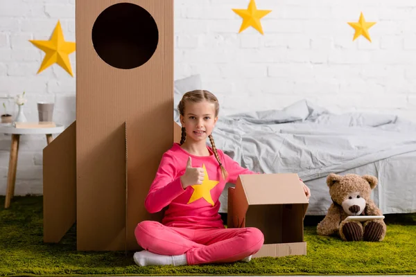 Smiling kid with braids sitting on carpet and showing thumb up in bedroom — Stock Photo