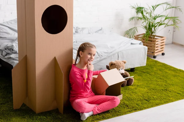 Curious kid in pink clothes sitting on carpet near cardboard rocket — Stock Photo
