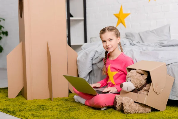 Smiling kid with teddy bear sitting on carpet and using laptop in bedroom — Stock Photo