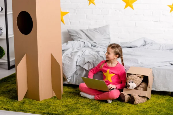 Niño sonriente sentado en una alfombra verde con portátil y oso de peluche - foto de stock