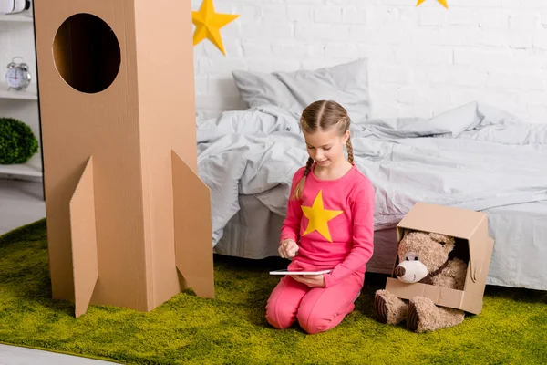 Cute kid with braids sitting on carpet and using digital tablet in bedroom — Stock Photo