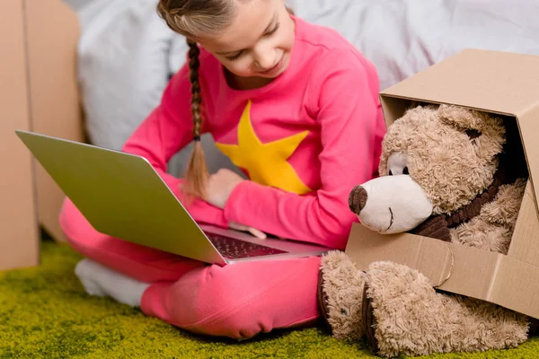 Cheerful kid with laptop sitting on carpet and looking at teddy bear — Stock Photo