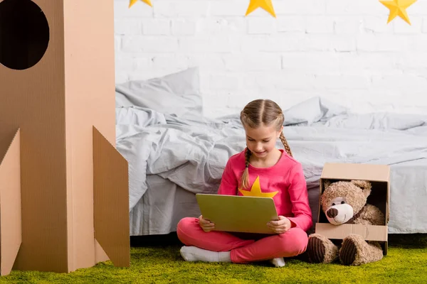 Enfant agréable assis sur le tapis avec ours en peluche et en utilisant un ordinateur portable — Photo de stock