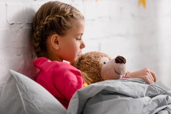 Vue latérale de mignon enfant couché dans le lit avec ours en peluche — Photo de stock