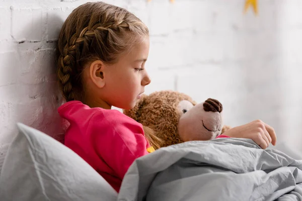Vista lateral de un niño lindo acostado en la cama con oso de peluche — Stock Photo