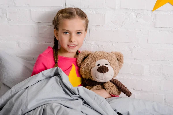 Neugieriges Kind mit Zöpfen hält Teddybär im Bett — Stockfoto