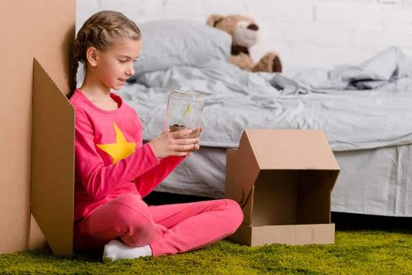 Interested kid sitting on carpet and holding plant in glass jar — Stock Photo