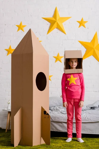 Enfant souriant dans un casque debout près d'une fusée en carton dans la chambre — Photo de stock