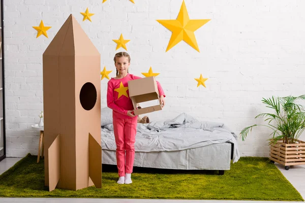 Cute child with cardboard helmet standing near rocket in bedroom — Stock Photo